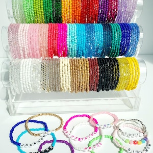 May include: A display of colorful beaded bracelets in various colors, including green, orange, red, and purple. Some bracelets have names spelled out with letter beads. The bracelets are arranged on clear acrylic stands, showcasing a variety of styles and designs.