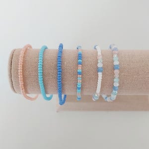 May include: A display of six beaded bracelets in shades of pink, blue, and white. The bracelets are strung on elastic and are of varying bead sizes. The bracelets are displayed on a tan fabric-covered cylinder.