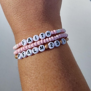 May include: Three beaded bracelets with the words "FAITH" and "PSALM 23" in white letters on pink and gold beads.