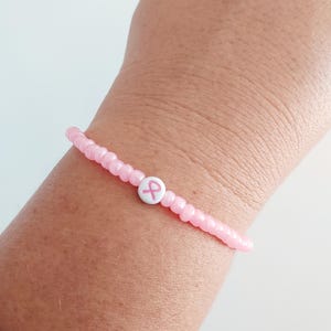 May include: Pink beaded bracelet with a white bead featuring a pink awareness ribbon. The bracelet is a delicate accessory, perfect for showing support. The beads are small and round, creating a simple yet meaningful design.