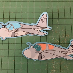 Puede incluir: Dos pegatinas de un avión militar de estilo caricatura. Los aviones son de color gris claro con detalles en azul y naranja. Una pegatina tiene las letras "NE" en la cola, y la otra un gráfico de delfín. El número "500" es visible en la nariz de cada avión.