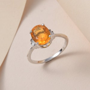 May include: A silver ring with an oval-shaped orange gemstone set in a delicate, intricate design. The ring has two smaller gemstones on either side of the main stone.