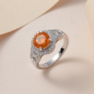 May include: A silver ring with a round orange gemstone surrounded by smaller white gemstones. The ring has a delicate design with a knot-like detail.