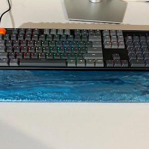 May include: A black mechanical keyboard with gray keycaps and colorful LED backlighting sits above a blue resin wrist rest. The wrist rest has a marbled ocean wave design. An orange key is visible.