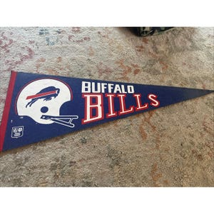 May include: A blue felt pennant with the words "BUFFALO BILLS" in white and red lettering. The pennant features the team's logo, a white helmet with a red charging buffalo. The pennant has a red border on the left side.