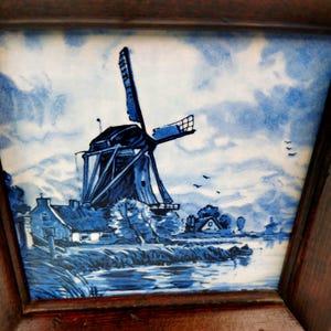 May include: A square ceramic tile featuring a blue and white hand-painted scene of a Dutch windmill situated by a river. The artwork depicts a traditional mill structure, a small house, and birds in the sky, all encased in a dark wooden frame.