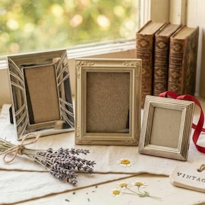 May include: Three rectangular picture frames of varying sizes and designs. One frame is silver with angled lines, one is cream-colored with ornate edges, and one is a smaller, simple silver frame with a red ribbon attached to the side.