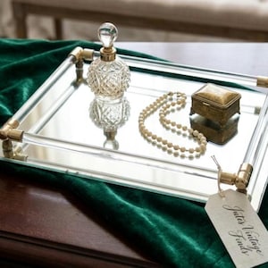 May include: A rectangular mirrored tray with clear acrylic side rails and gold-tone metal accents. On the tray sits a clear glass perfume bottle with a diamond-cut pattern, a string of cream-colored pearls, and a small gold-tone metal box with a tag.