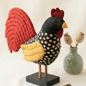 May include: A fabric rooster figurine featuring a black body with cream-colored polka dots, a large red quilted tail, and yellow quilted wings. The rooster has a red comb and wattle, a yellow beak, and stands on two wooden legs attached to a black base.