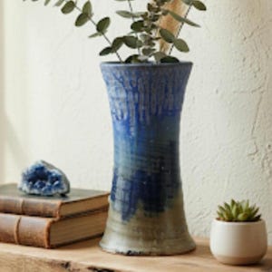 May include: A tall, blue and green ceramic vase with a textured surface. The vase is filled with green leafy branches. A blue geode, two leather-bound books, and a small potted succulent are also visible.