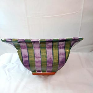 May include: A square ceramic bowl featuring vertical stripes in alternating shades of purple and olive green. The bowl has a wide, flared rim and a solid orange base, with a glossy finish that covers the exterior surface of the vessel.