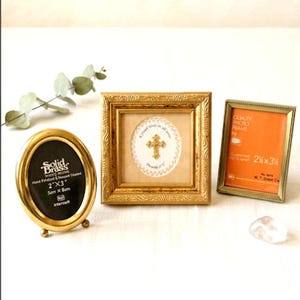 May include: Three small picture frames: an oval solid brass frame labeled 2"x3", a square gold frame with an ornate border containing a cross illustration with the text "A friend loves at all times Proverbs 17:17", and a rectangular gold frame.
