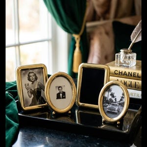May include: A collection of gold-toned picture frames in various shapes, including rectangular and oval, displayed on a black tray. The frames hold black and white photographs of a woman, a man, and a vintage car. A glass inkwell and books are in the background.