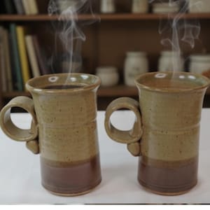 May include: Two handmade ceramic mugs with a unique handle design. The mugs are filled with a dark liquid, likely coffee, and steam rises from them. The mugs have a two-tone color scheme, with a light brown upper section and a darker brown base.