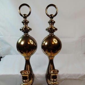 May include: Two ornate, gold-colored candle holders. Each has a ring at the top, a spherical center, and a candle cup at the bottom. The design is symmetrical and features a polished, reflective surface.