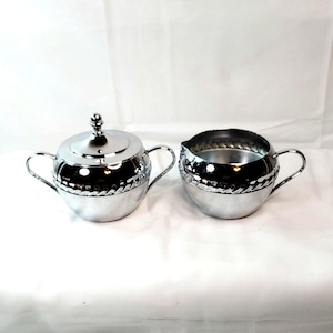 May include: A polished silver-tone metal sugar bowl with a matching lid and a creamer pitcher. Both pieces feature two curved handles and a decorative rope-style embossed band around the upper section, reflecting light on their smooth surfaces.