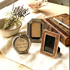 May include: A silver apple-shaped frame with the text With sincere thanks and best wishes, a silver octagonal frame, and a rectangular brown marbled frame. These three frames are arranged alongside a bowl of dried lavender and a vintage book.