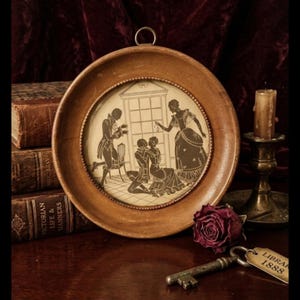 May include: A framed silhouette art piece depicting a scene with figures in period clothing. The round wooden frame has a brass hanging loop. A key and a tag that reads "LIBRA 1888" are in the foreground. A candle and books are in the background.