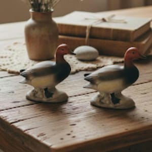May include: Two ceramic duck figurines with red heads, gray bodies, and white bellies are displayed on a wooden table. The ducks are perched on a textured base. A vase with dried flowers and a stack of books are in the background.
