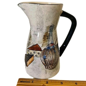 May include: A ceramic pitcher with a black handle and a spout. The pitcher is decorated with illustrations of cheese and a wine bottle. The pitcher measures approximately 6 inches tall.