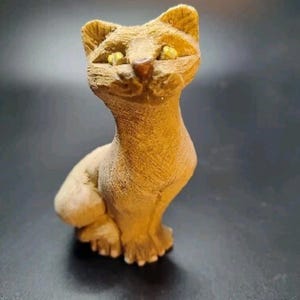 May include: A tan-colored cat figurine with yellow eyes and a detailed textured surface. The cat is in a seated position, with its front paws visible. The figurine has a slightly elongated neck and pointed ears.