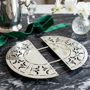 May include: Two-piece silver-toned serving dish with intricate cut-out designs. The dish is set on a dark, marbled surface, with a green ribbon, a silver ice bucket, and a small vase of white orchids in the background.