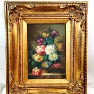 May include: An oil painting of a floral bouquet in a vase, featuring pink, white, yellow, and purple flowers with green leaves. The artwork is encased in a rectangular, ornate, metallic gold-colored frame with decorative corner carvings.