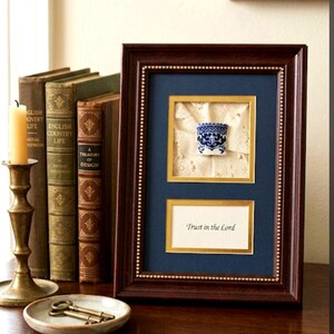May include: A dark brown wooden frame containing a blue and white ceramic thimble mounted on cream lace, positioned above a white rectangular insert with the black printed text Trust in the Lord, all set against a deep blue matting.