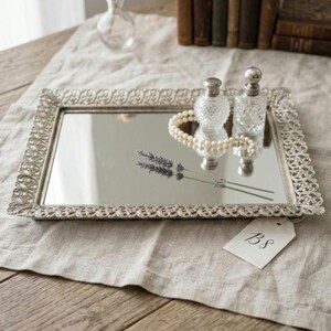 May include: A rectangular silver-toned mirrored tray featuring an intricate, ornate filigree border. Resting on the reflective surface are two clear glass perfume bottles, a strand of white pearls, and a sprig of lavender. A tag labeled 138 is attached.