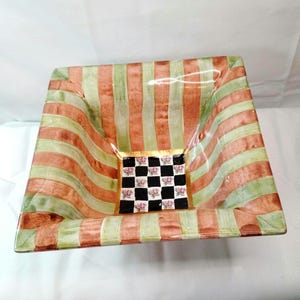 May include: A square ceramic bowl featuring alternating vertical stripes of muted green and terracotta orange. The center of the bowl displays a black and white checkerboard pattern with small pink floral accents, bordered by a thin gold line.