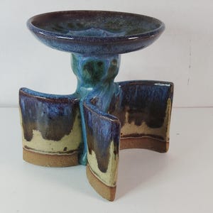 May include: A handcrafted ceramic pedestal bowl with a unique, abstract design. The bowl features a deep blue glaze with hints of green and brown. The base has three curved supports, and the bowl is approximately 6 inches in diameter.