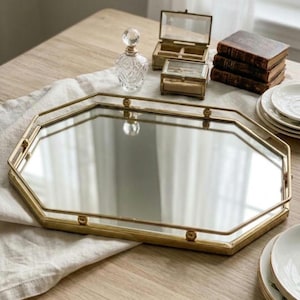 May include: An octagonal mirrored tray with a thin gold-toned metal frame and small spherical accents along the edges. The tray features a reflective glass surface and a raised border, providing a flat base for displaying various decorative items.