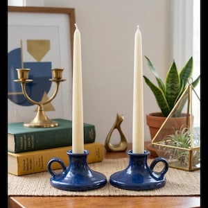 May include: Two blue ceramic candle holders with cream-colored taper candles. The candle holders have a handle on the side. The background includes books, a brass candelabra, and a potted plant.