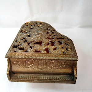 May include: A gold-toned metal trinket box shaped like a grand piano. The lid features an intricate openwork floral filigree design with a red interior visible beneath. The sides are decorated with embossed musical lyre motifs and floral patterns.