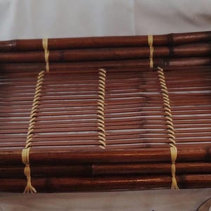 May include: A rectangular bamboo serving tray with a dark brown finish. The tray is constructed from bamboo poles and slats, with light-colored twine used to secure the slats and frame. The tray has a natural, rustic aesthetic.
