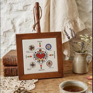 May include: A square wooden trivet with a ceramic tile insert. The tile features a central heart design with the words "Bless this House" and surrounding floral and geometric patterns. A wooden handle with a leather loop is attached.
