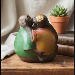 May include: A ceramic sculpture depicts two figures embracing and kissing. One figure wears a green top, the other a yellow and pink top. The sculpture is on a wooden surface, with a small potted succulent and books in the background.