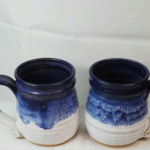 May include: Two handmade ceramic mugs with a blue and white glaze. The mugs have a dark blue rim and handle, with a white base. The blue glaze fades into the white, creating a watercolor effect. These mugs are perfect for coffee or tea.