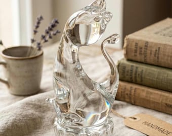 May include: A clear glass figurine of a cat in a seated position with its tail curved upward. The cat is perched on a circular, textured base with vertical ridges. The glass is transparent and reflects light, showing smooth, rounded contours.