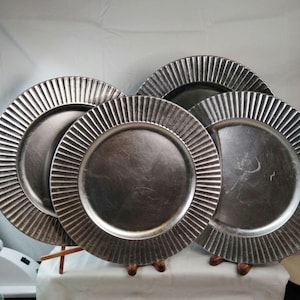 May include: A set of three round silver-toned charger plates with a metallic finish. Each plate features a wide, textured rim with vertical fluted grooves and a smooth, circular center. The plates are arranged in an overlapping display.