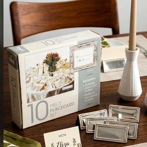 May include: A 10-piece place card set in a rectangular box with a photo of a table setting. The set includes silver-toned rectangular place card holders with a raised border. One holder displays a card with the name Eliza and Table 4 printed.