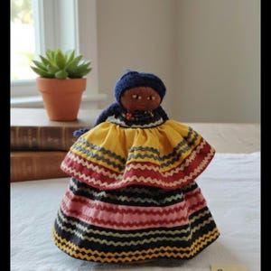 May include: A handmade doll with a dark complexion, wearing a blue cap and a colorful dress. The dress features layers of yellow, red, pink, and black fabric with zigzag patterns. The doll is a folk art item.