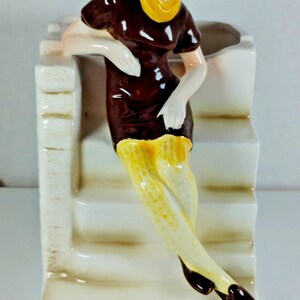 May include: A ceramic figurine of a woman in a brown dress with a yellow collar and patterned stockings, leaning against a white structure. The figurine has brown hair and black shoes. The base is white with a staircase design.