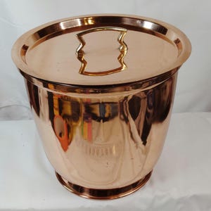 May include: A copper ice bucket with a matching lid. The lid has a gold-colored handle. The bucket has a rounded shape and a shiny finish. The bucket is designed to hold ice and keep beverages cold.