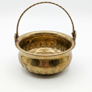 May include: A small, round, metallic brass bowl with a polished finish and a textured, repeating pattern along the inner and outer rim. The bowl features a thin, twisted metal handle attached to two small side loops, allowing it to swing freely.