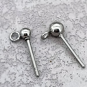 Puede incluir: Dos piezas de joyería corporal plateadas estilo barra. Cada pieza presenta una pequeña bola redonda en un extremo y un poste recto y delgado. Una tiene un pequeño lazo adjunto a la bola. La joyería se muestra sobre una superficie gris clara texturizada.