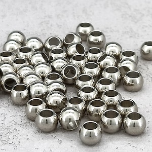 May include: A pile of silver metal beads, each with a small hole in the center. The beads are round and smooth, and they are scattered on a light gray surface.