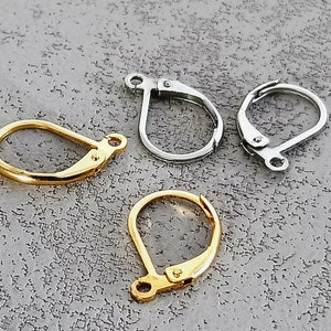 Puede incluir: Primer plano de cuatro cierres de pendientes de palanca. Dos son de color dorado y dos de color plateado. Los cierres están sobre una superficie gris texturizada. Los cierres de palanca se utilizan para asegurar los pendientes.