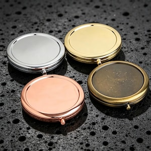 Op de afbeelding: Vier ronde compacte spiegels in verschillende metalen afwerkingen, waaronder zilver, gepolijst goud, roségoud en een antieke messingkleur. Elke ronde behuizing heeft een glad, reflecterend oppervlak en een klein scharniermechanisme.