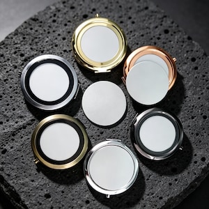 May include: A collection of six round compact mirrors in various metallic finishes including gold, silver, rose gold, and black, arranged around a central plain white circular disc. Each mirror features a polished frame and a reflective surface.